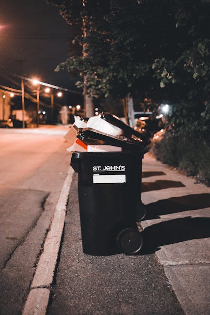A black wheeled rubbish bin positioned on a pavement beside a road at night, with a white label and the words 'ST. JOHN'S' printed on the front. The lid of the bin is open, revealing a mixture of card